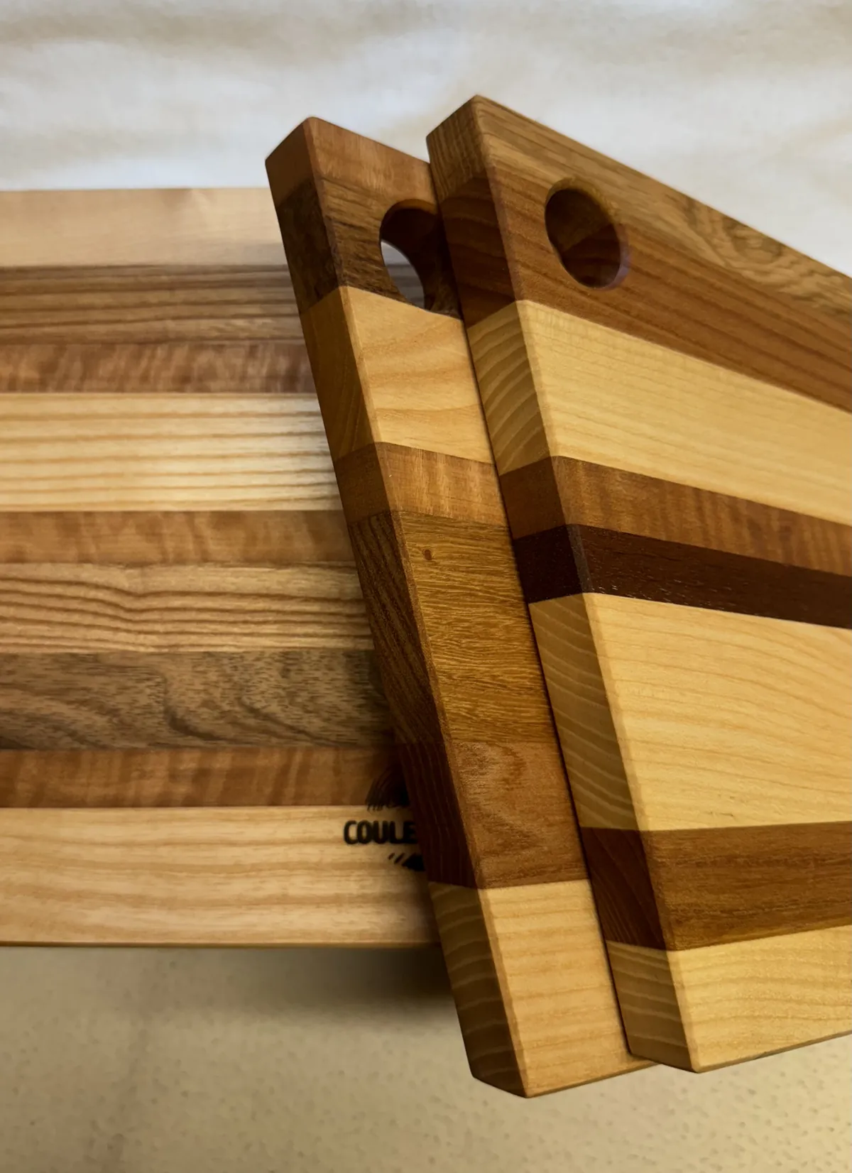 Deux planches à découper en bois massif, bandes de frêne, noyer et cerisier, percées d'un trou de suspension, posées sur une planche gravée CouleurBois